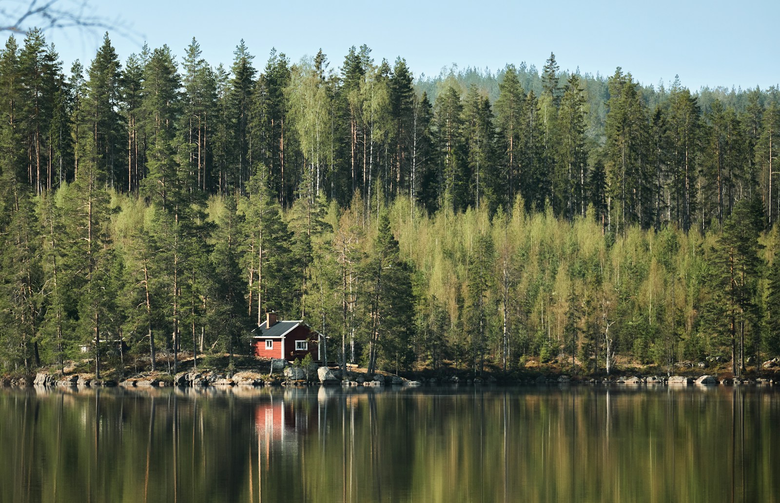 A red cottage reflected in a quiet lake, with forest behind it.